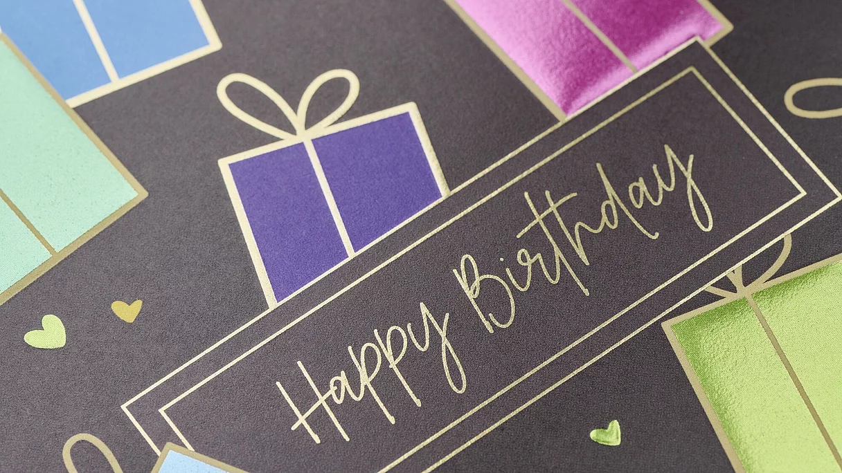  close-up of a printed card with "Happy Birthday" written in a delicate, handwritten-style font. The card features a dark background adorned with colorful, shiny presents and small hearts.