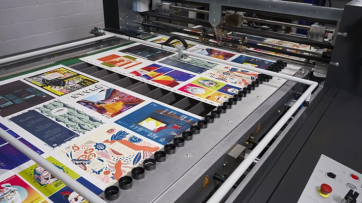 Printing machine producing vibrant, colorful sheets with various illustrated designs and patterns on a conveyor belt.