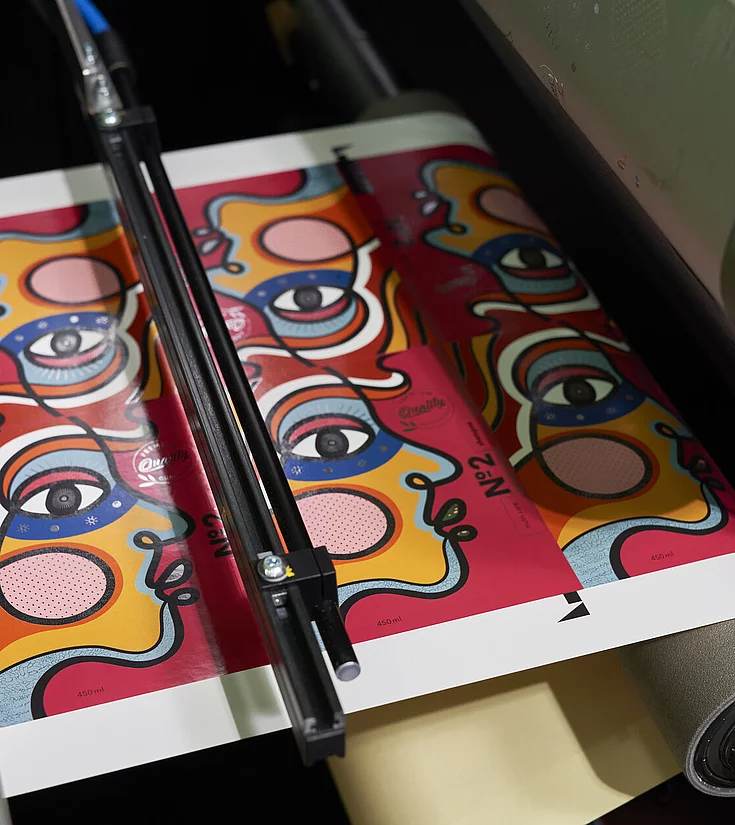 Printed sheet with colorful abstract face illustrations undergoing foil embellishment in a KURZ digital embellishment system.