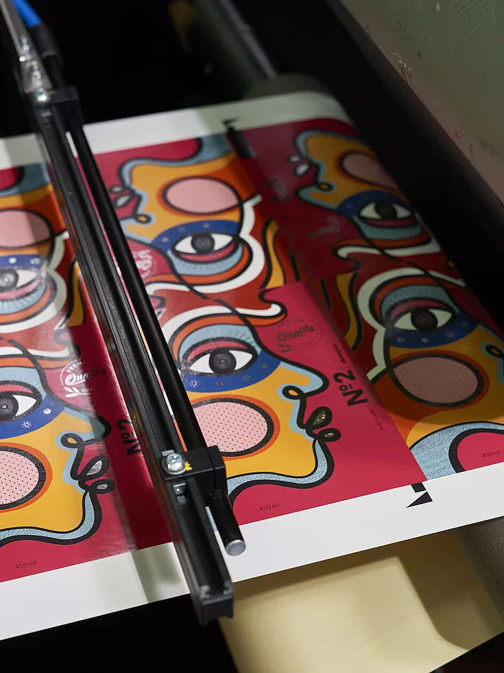 Printed sheet with colorful face illustrations being foil embellished in a KURZ digital system.