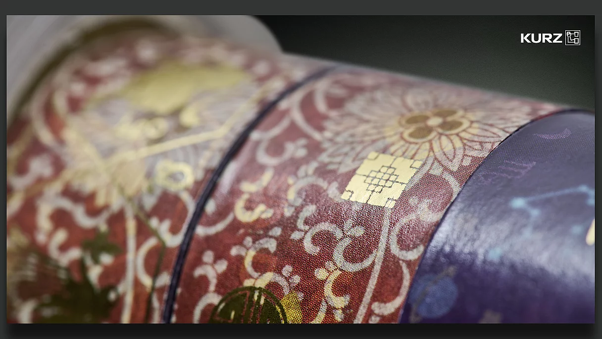 Close-up of a textured, patterned packaging with red and purple colors and gold foil accents, featuring floral and decorative designs and the KURZ logo.