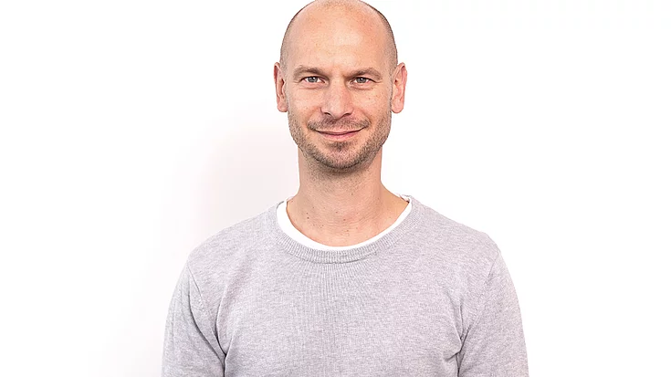 Smiling bald man with light facial hair wearing a light gray sweater, standing against a white background.