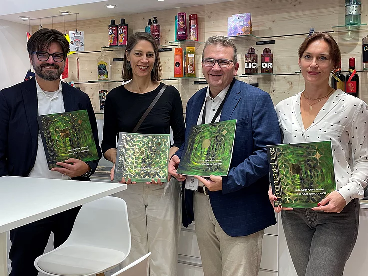Four people standing indoors holding copies of Formes de Luxe magazine, smiling at the camera, with shelves of colorful bottles and packaging in the background.