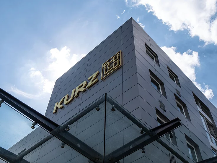Modern KURZ company building with metallic facade and gold logo, photographed from below against a blue sky with clouds.