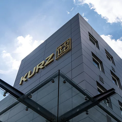 Modern KURZ company building with metallic facade and gold logo, photographed from below against a blue sky with clouds.