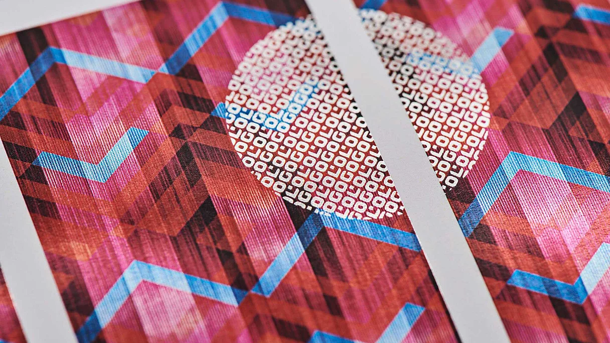 Nahaufnahme eines bedruckten Blattes mit geometrischem Muster in Rot-, Rosa- und Blautönen, auf dem ein weißer Kreis mit kleinen quadratischen Formen zu sehen ist, der durch einen weißen vertikalen Streifen unterteilt ist.