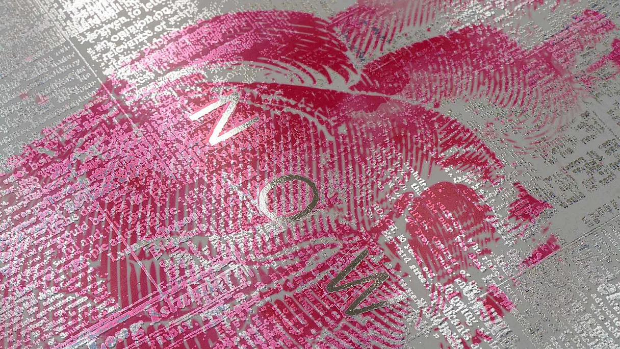 Nahaufnahme eines bedruckten Blattes mit silbernem Folientext und einem darüber liegenden rosa Lippenstiftabdruck. In der Mitte des Bildes ist das Wort „NOW“ zu sehen.