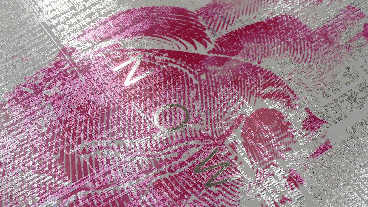 Close-up of a printed sheet with silver foil text and a pink lipstick print overlaid. The word "NOW" is visible in the middle of the image.