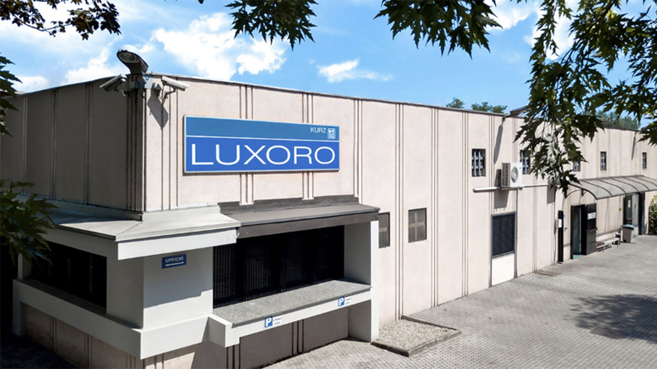 Exterior view of a beige industrial building with a blue sign reading ‘LUXORO’ mounted above the entrance, surrounded by trees and a paved driveway