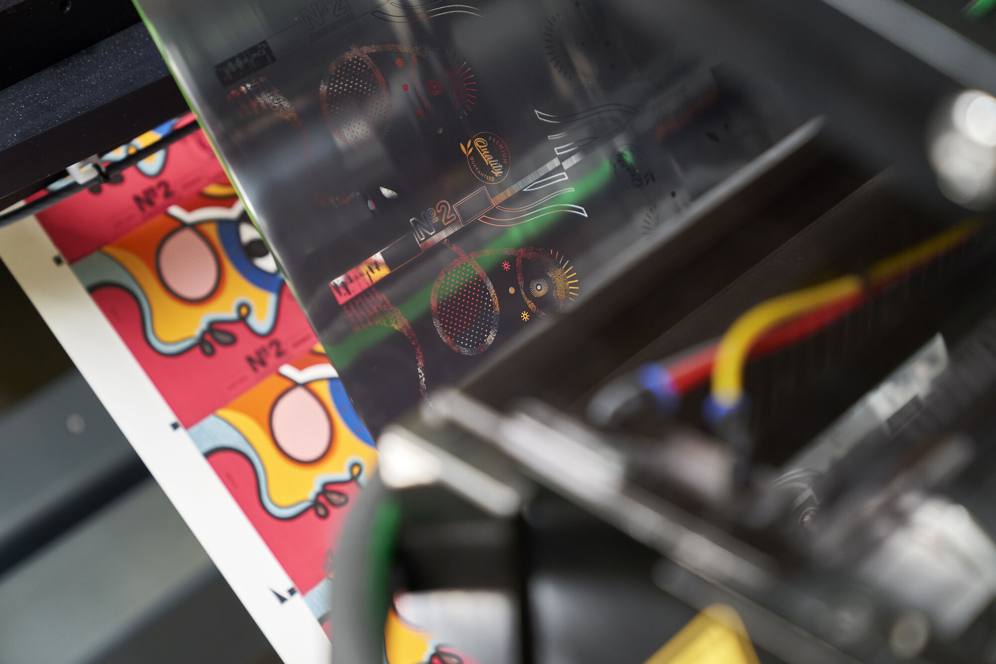 Close-up of KURZ digital embellishment machine applying holographic foil to colorful printed sheet with abstract artwork and product label design.